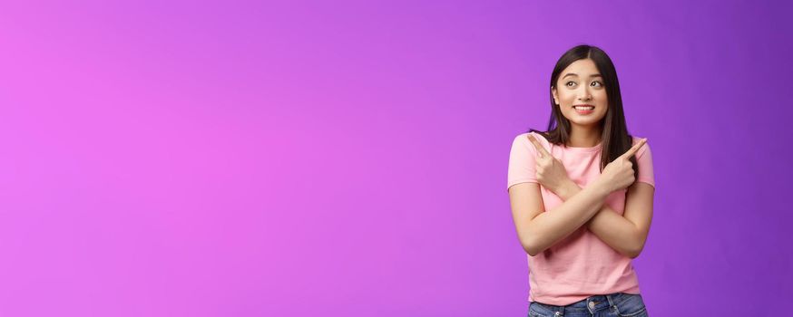 Cheerful timid cute asian girl, look tender, smiling gazing aside cross hands chest, pointing sideways, show left right, making decision, gathering confidence choose, purple background.