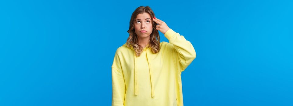 Gosh just kill me. Bored and pressured, stressed young blonde girl in yellow hoodie, make pistol gesture near head, shooting from gun and sighing as feel bothered and troubled, blue background.