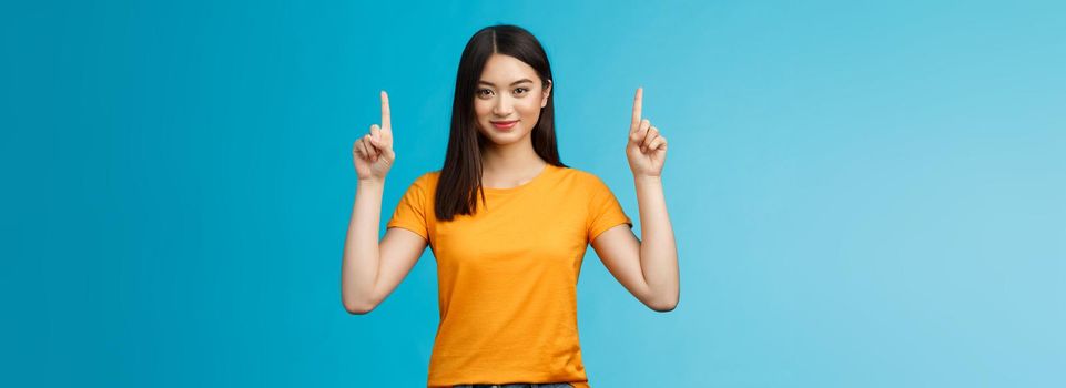 Heads up see awesome promo. Assertive good-looking confident asian woman showing friend advertisement, raise index fingers pointing top, smiling camera self-assured, stand blue background determined.