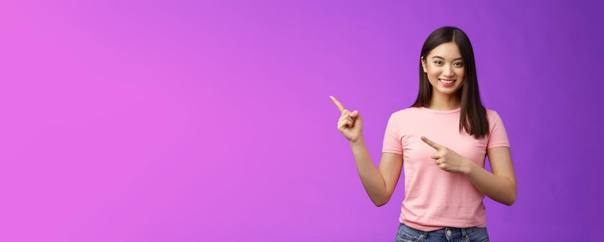 Friendly cute pleasant asian woman showing way, indicating interesting promo, pointing left smiling broadly, introduce advertisement product, stand purple background cheerful. Copy space