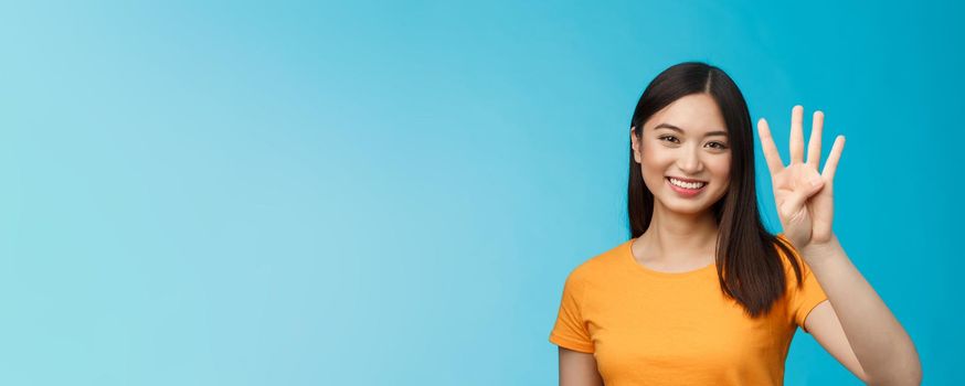 Close-up charismatic tender stylish asian female show number four fingers, smiling happily, making order, explain how much items want buy, stand blue background carefree. Copy space