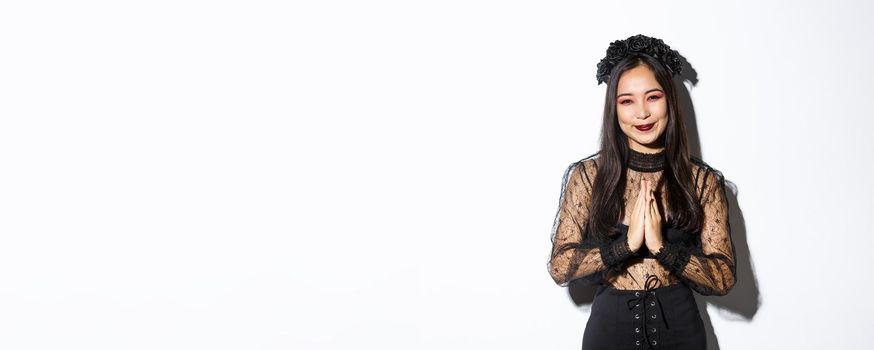 Image of happy and grateful young asian woman in gothic lace dress clasp hands together to say thank you, smiling thankful and standing over white background.