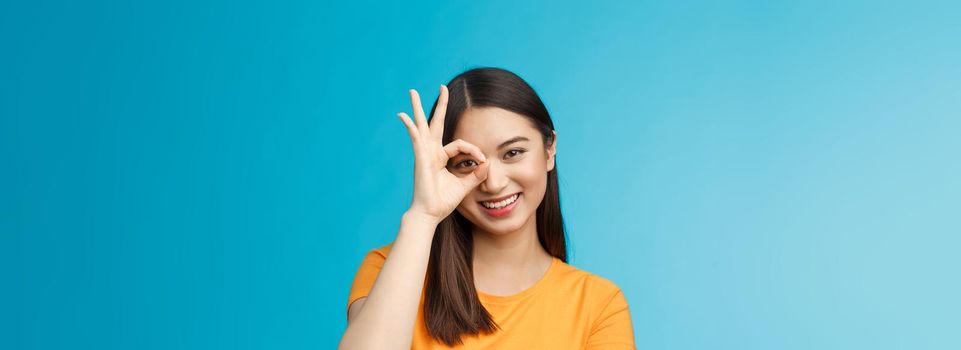 Close-up cute dreamy tender asian woman dark short haircut show okay ok sign look through circle smiling broadly, express positivity good mood, standing upbeat friendly gaze camera, blue background.