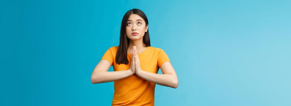 Serious-looking hopeful cute asian woman begging god hear prayers, supplicating, making wish look up sky talking lors, believe have faith, stand blue background pleading good fortune luck.