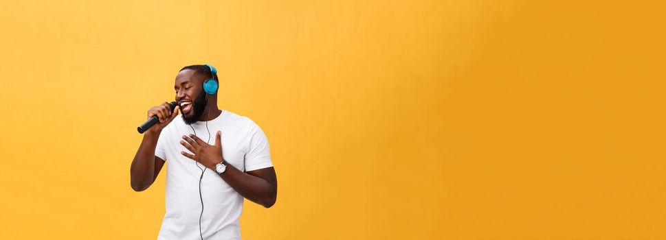 Portrait of cheerful positive chic. handsome african man holding microphone and having headphones on head listening music singing song enjoying weekend vacation isolated on yellow background.