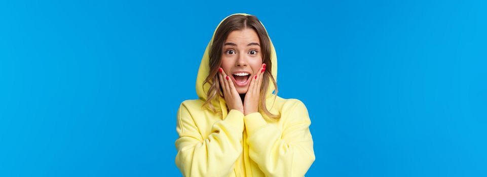 Close-up portrait of impressed and fascinated blond girl in hood react to amazing great news, touch cheeks gasping and smiling wondered, hear she won prize, standing blue background.