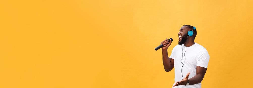 Portrait of cheerful positive chic. handsome african man holding microphone and having headphones on head listening music singing song enjoying weekend vacation isolated on yellow background.