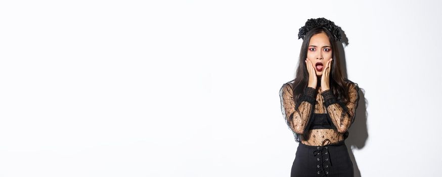 Shocked young asian woman wearing witch costume, looking concerned. Female in black dress and wreath looking like widow or magician gasping worried, standing white background.