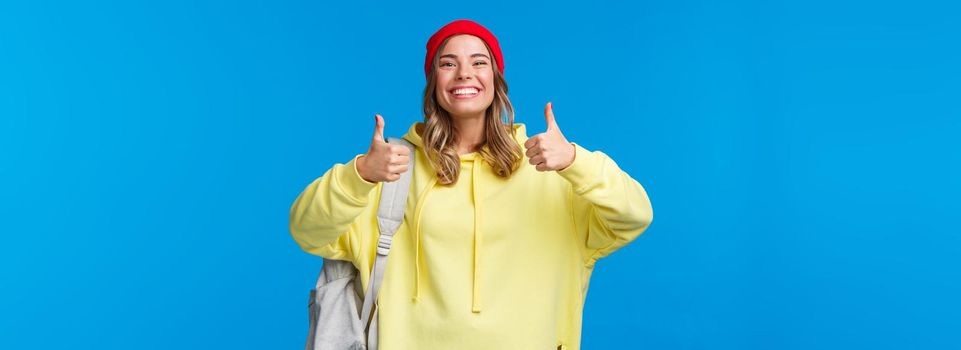 Sounds good, deal. Cheerful happy and smiling hipster girl in red beanie and yellow hoodie, approve idea, agree with friends picking where hang out after classes, hold backpack, blue background.