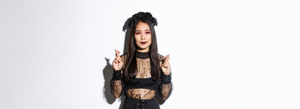 Image of hopeful asian girl in gothic lace dress and wreath making wish, cross fingers for good luck, looking wishful at camera, standing over white background.