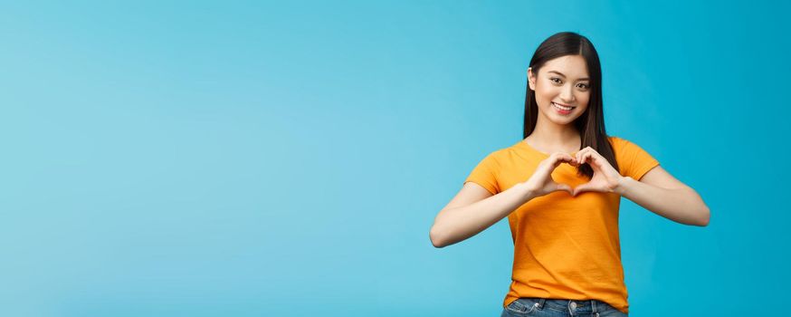 Lovely caring attractive asian girlfriend show heart sign cherish and value relationship, smiling broadly supportive, stand blue background wear yellow t-shirt, promote peace and happiness.