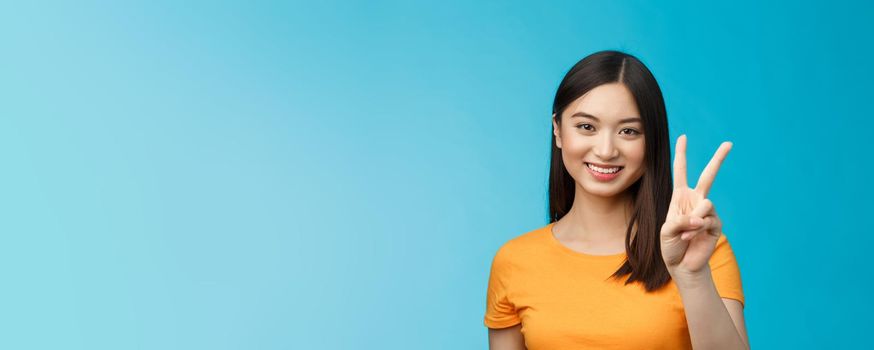 Cheerful friendly asian woman in yellow t-shirt smiling broadly, show number two, twice, win second place, smiling joyfully, make take-away order, book sits, stand blue background.
