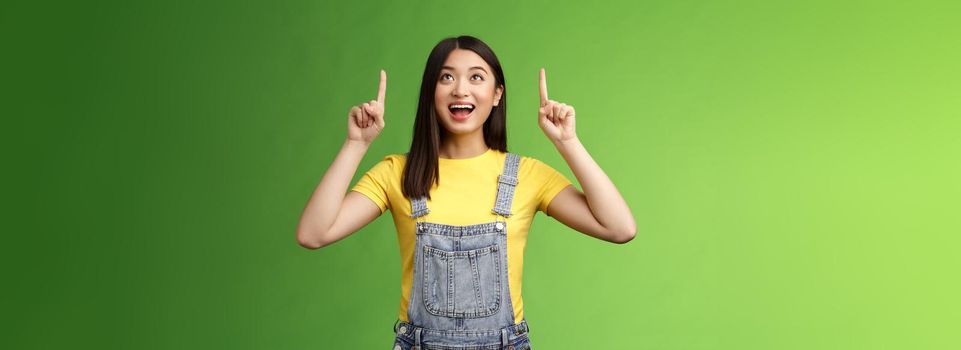 Carefree amused asian brunette in yellow t-shirt dungarees, smiling amazed and fascinated, look pointing up, observe cool new promo, grinning excited, enjoy watching top copy space.