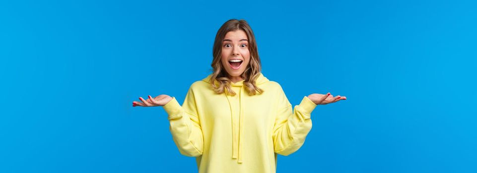 What nice surprise. Amused and happy cute blond caucasian woman spread hands sideways and shrugging smiling with excitement and disbelief, hear unexpected good news, blue background.