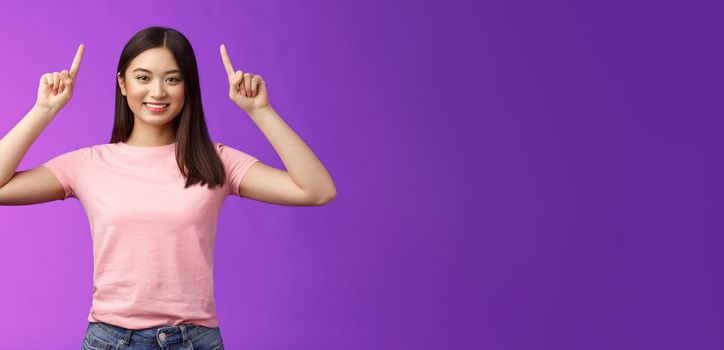 Cute assertive dark-haired asian woman wear pink t-shirt raise hands up, showing promo, pointing fingers up, grinning, inviting use link, try-out product, give direction, stand purple background.