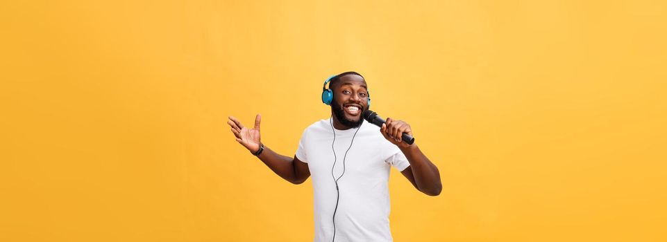 Portrait of cheerful positive chic. handsome african man holding microphone and having headphones on head listening music singing song enjoying weekend vacation isolated on yellow background.