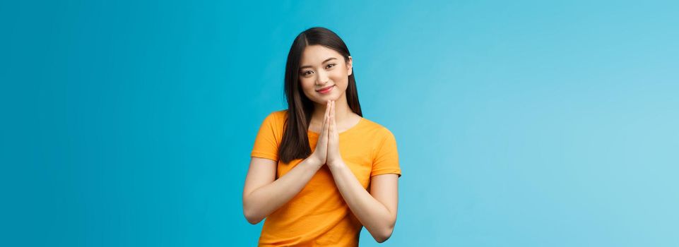 Pretty please girl asking politely. Cute charming asian woman smiling tenderly, hold hands pray grinning begging for favour, plead, look thankful for help stand blue background joyful, caring.