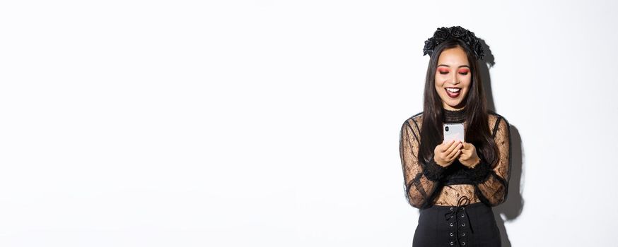 Excited smiling woman looking at mobile phone pleased, wearing gothic lace dress for halloween party, standing over white background.