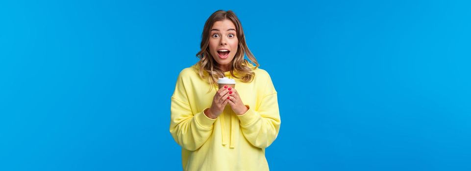 Surprised cute blond caucasian girl react to amazing news or awesome promo in her favorite cafe, holding take-away cup of coffee gasping and look camera fascinated, blue background.