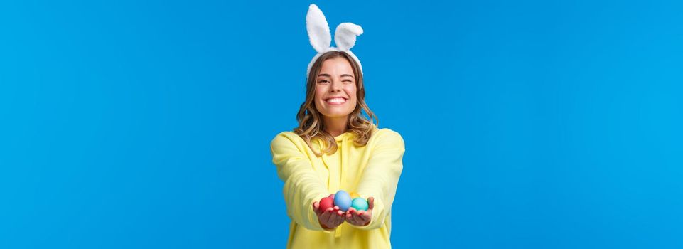 Holidays, people and emotions concept. Tender cute blond caucasian female giving Easter eggs to you with beaming pleasant smile, wearing rabbit ears, celebrating religious holiday.