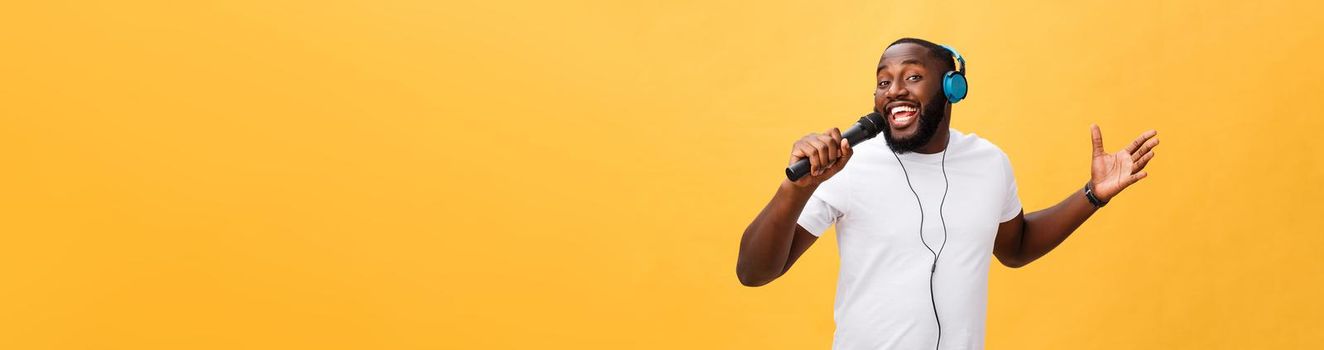 Portrait of cheerful positive chic. handsome african man holding microphone and having headphones on head listening music singing song enjoying weekend vacation isolated on yellow background.