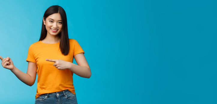 Friendly outgoing good-looking asian woman showing promo, pointing left indicating fingers advertisement, give advice, smiling confident, pleasantly help pick clothes, stand blue background.