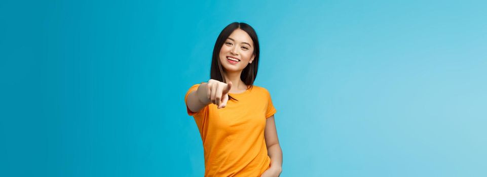 Lucky winner. Cheerful charismatic asian cute urban girl stand yellow t-shirt smiling friendly pointing finger camera choosing, picking person, inviting you team, stand happy blue background.