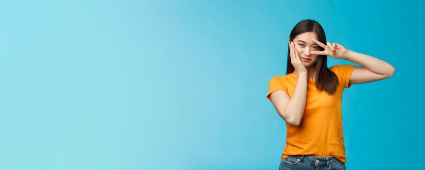 Sassy stylish good-looking asian assertive korean girl show peace victory sign on eye, touch cheek look empowered and motivated, standing cheeky coquettish blue background, wear yellow t-shirt.