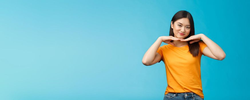 Sweet lovely tender asian girl brunette, tilt head smiling show clean pure face hold palms under jawline satisfied after applying skincare product stand blue background wear yellow t-shirt.