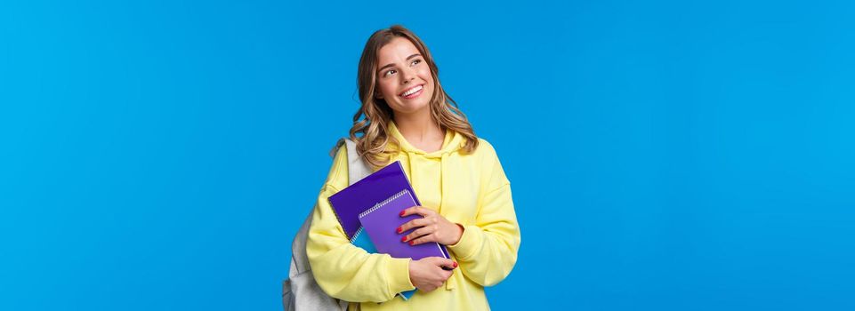 Dreamy happy good-looking pretty blond girl studying in college, holding backpack and notebooks, homework staff, look upper left corner thoughtful, standing blue background.