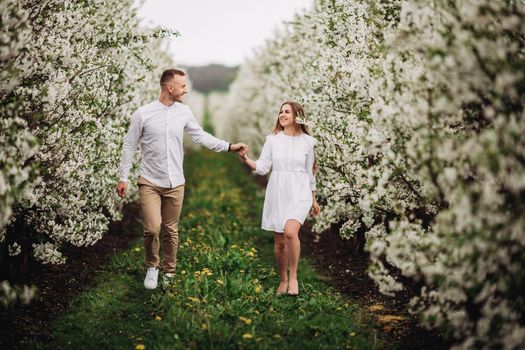 Happy family couple in spring blooming apple orchard. Young couple in love enjoy each other while walking in the garden. The man holds the woman's hand. Family relationships