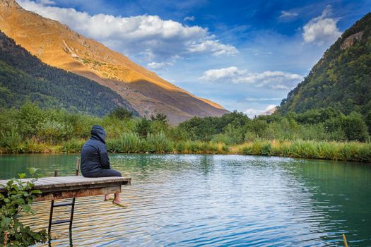 A man sits on a bridge near a mountain lake, enjoying the beauty of the mountains