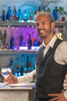 Attractive young barman in a white shirt and black apron smiling looking to camera at party in nightclub. Night lifestyle concept. High quality photo