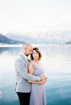 Groom hugs bride from behind by the sea with a view of the mountains. High quality photo