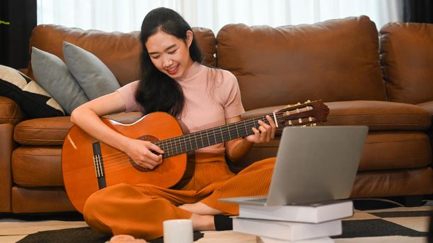 Smiling young woman learning play guitar, watching online lessons on laptop. Hobbies and learning concept.