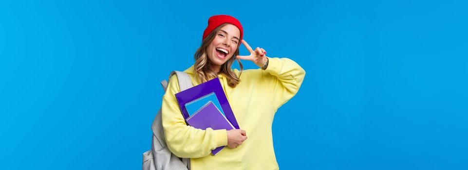Education and learning concept. Cheerful smiling pretty female student showing peace gesture, enjoying college life, wink and gaze optimistic, studying hard, holding notebooks and backpack.