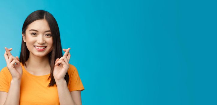 Close-up hopeful cute asian girl with short dark hair cross fingers good luck, praying smiling broadly awaiting positive news, faithfully hope win, stand blue background upbeat excited.