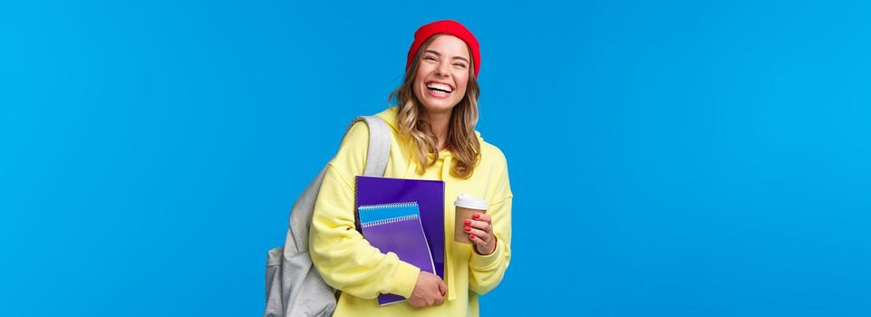Cheerful happy female student having fun, enjoying university lifestyle, carry backpack and notebooks, drinking take-away coffee, talking to classmates, standing blue background joyful.