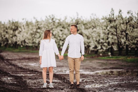 Happy family couple in the spring blooming apple orchard. Young couple in love enjoy each other while walking in the garden. Mud underfoot. The man holds the woman's hand. Family relationships