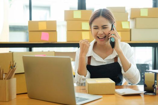 Female small online business owner talking on phone with clients with excitement and delight to recieve and check order online, address preparing to pack order boxes for delivery. Online business concept.