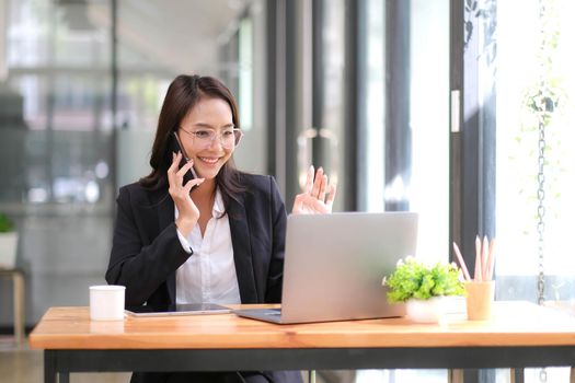 Happy businesswoman sitting at desk behind her laptop and talking with somebody on her mobile phone while working at office..