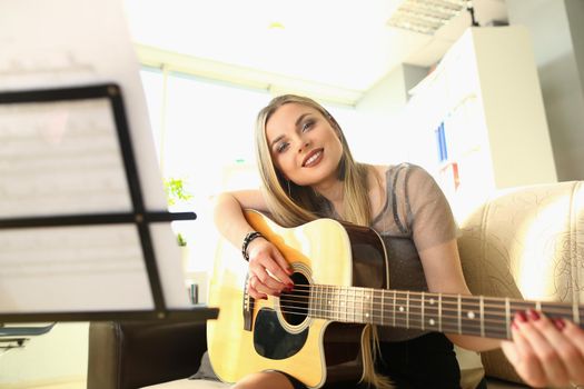 Smiling blond woman playing guitar and looking through notes. Talented musician and music hobby concept