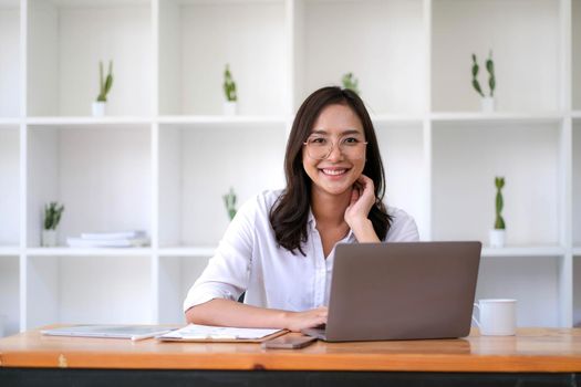 Smiling beautiful young Asian businesswoman sitting with laptop and computer while doing some paperwork at the office. Looking at camera..