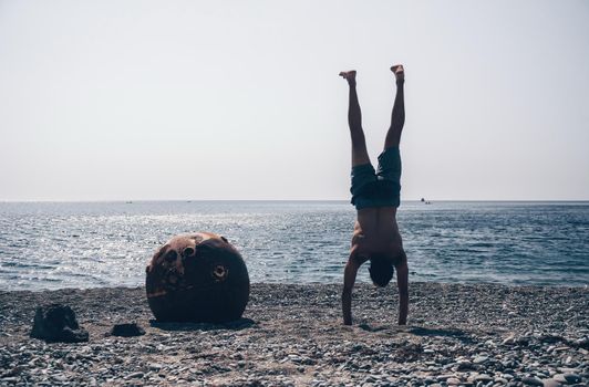 Man doing yoga poses standing on old rusty floating marine mine on the beach with rocky shore and sea background