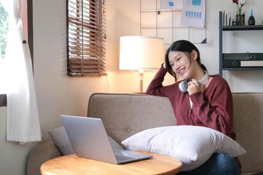 Beautiful Asian woman using laptop while headphones sitting on sofa at home.