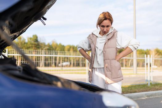 a young woman at the open hood of a car is trying to figure out the malfunction of the car.