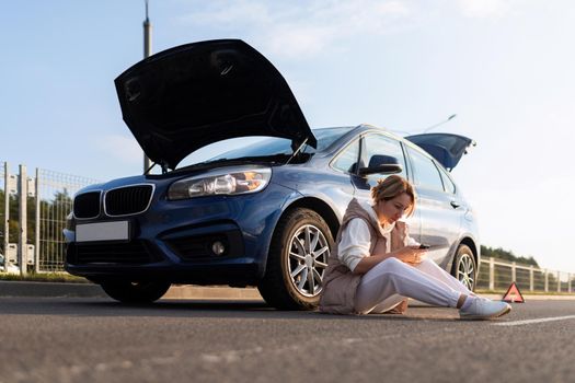 frustrated woman sitting on the road near the broken down car with the hood up.