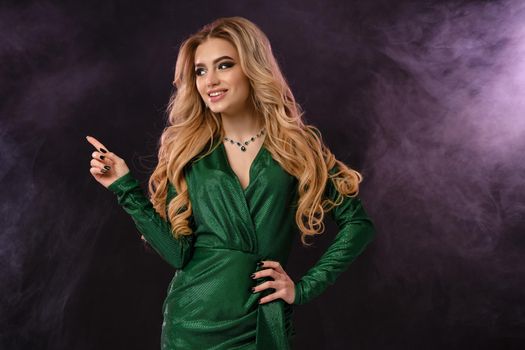 Gorgeous blonde female, make-up, in green dress and jewelry. Smiling, pointing at something, posing on black smoky background. Template, mockup for your advertising and design. Close up, copy space
