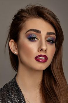 Brunette lady in black shiny dress is looking at you, posing on gray background. Luxury makeup, perfect skin. Colorful eyeshadow, false eyelashes, glossy red lips. Professional maquillage. Close up