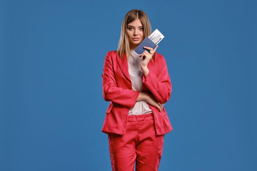 Alluring blonde lady in white blouse and red pantsuit. She has folded her hands, holding passport and ticket, posing against blue studio background. Travelling concept. Close-up, copy space